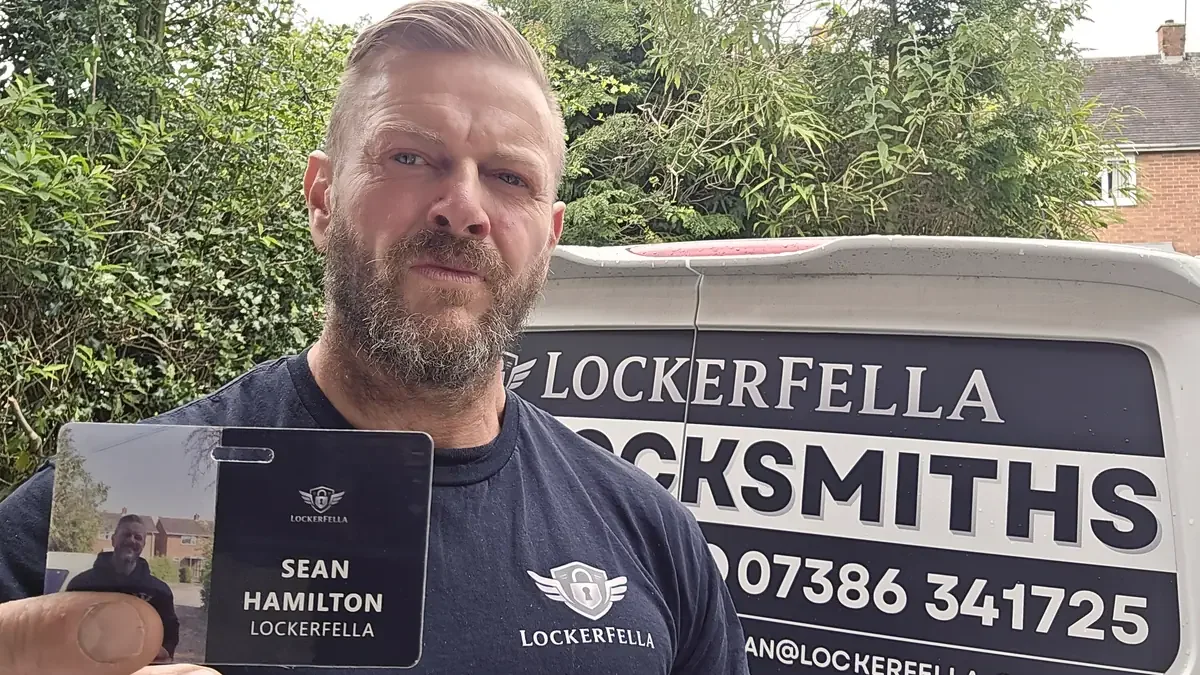 Sean Hamilton, the owner-operator of Lockerfella, photographed holding a printed identification card showing his name, role and the Lockerfella logo. He is wearing the same branded T-shirt visible in the featured image. The identification card is the visible artefact that turns the abstract claim 'I am the locksmith' into something a customer can read at the door before opening it.